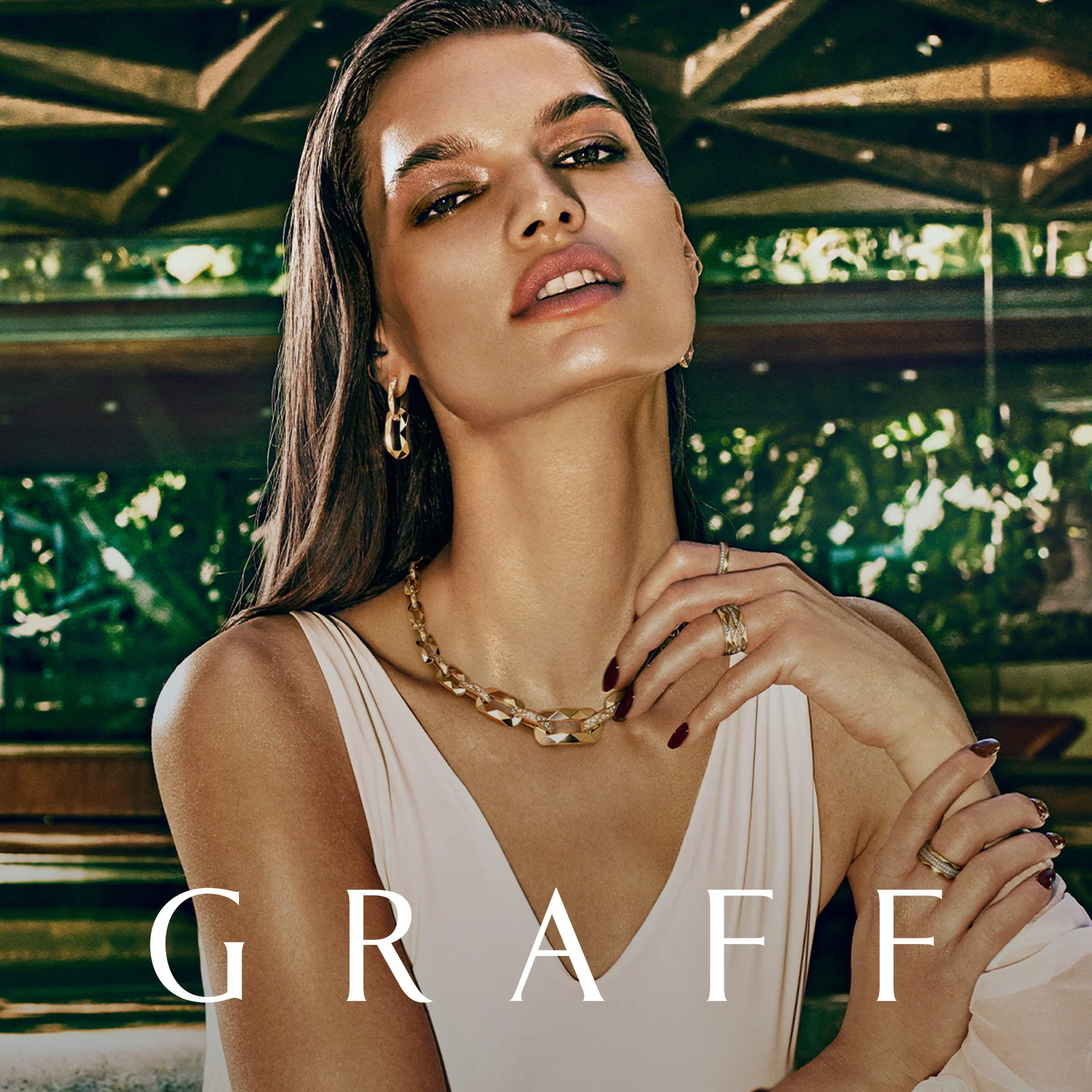 Woman wearing a white sleeveless top and gold and diamond necklace, earrings and rings by Graff Diamonds. Text 'GRAFF'. 