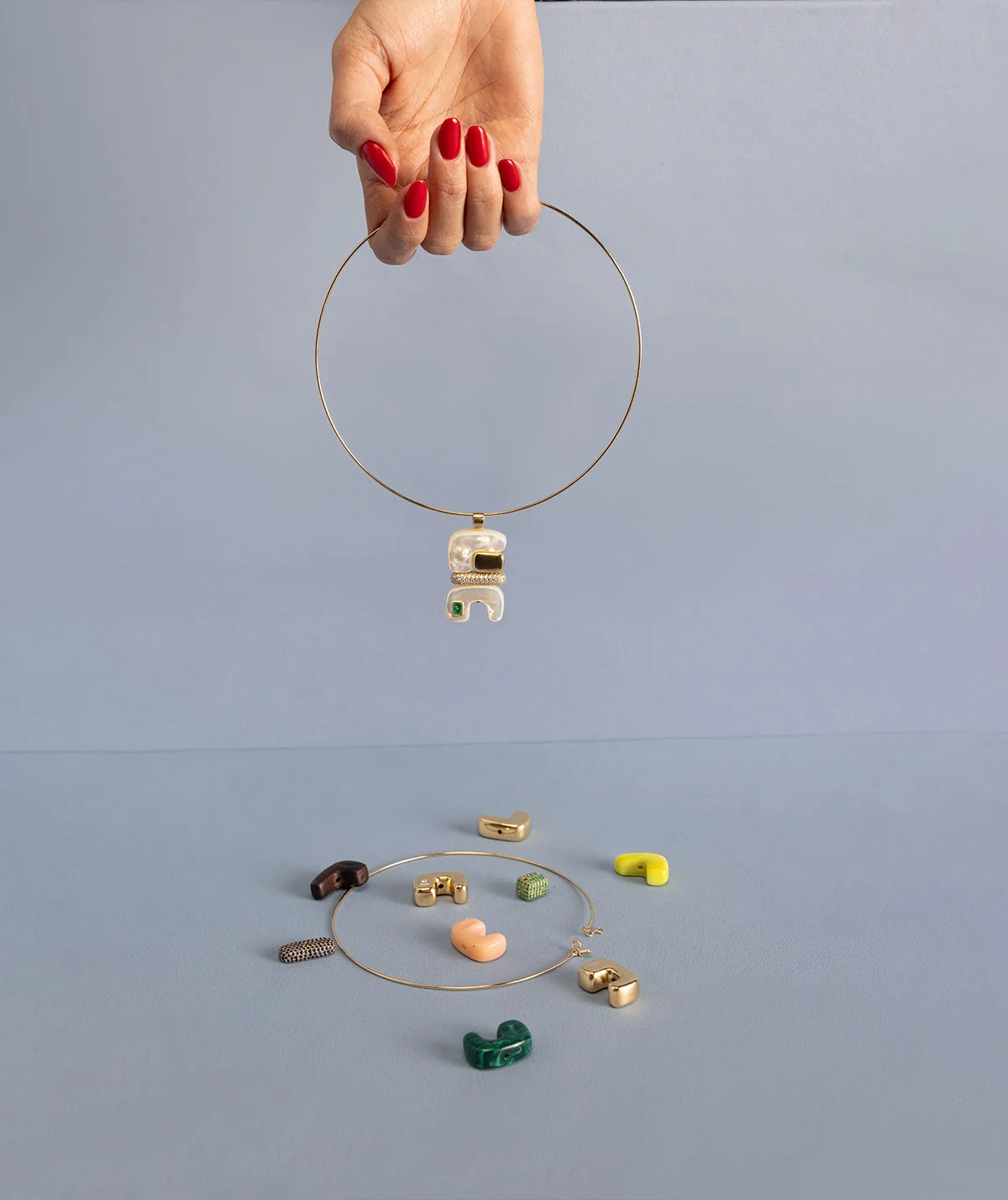 A hand with red nail polish holds a thin gold hoop necklace with a Joelle Kharrat pendant featuring a white face. Below, several small colorful pendants are scattered on a light blue-gray surface. 