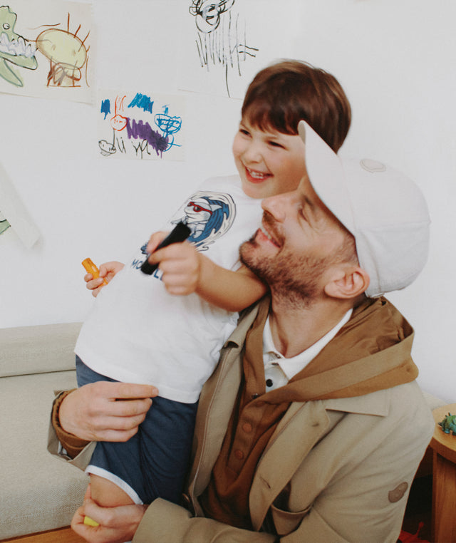 Man holding a child in a room with colorful drawings on the wall wearing Moncler jacket and baseball cap 
