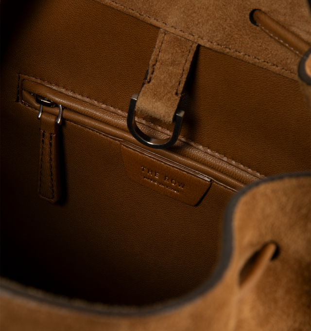 Image 3 of 3 - BROWN - THE ROW Backpack 9 in suede featuring a toggle fastening top, smooth leather trim, gold hardware, 2 adjustable, rivet fastening straps, 1 top handle, logo embossed purse feet at underside, single interior compartment with 1 slit pocket, key ring inside and suede lining. 10.5 W x 10.5 H. 100% calfskin leather. Made in Italy.