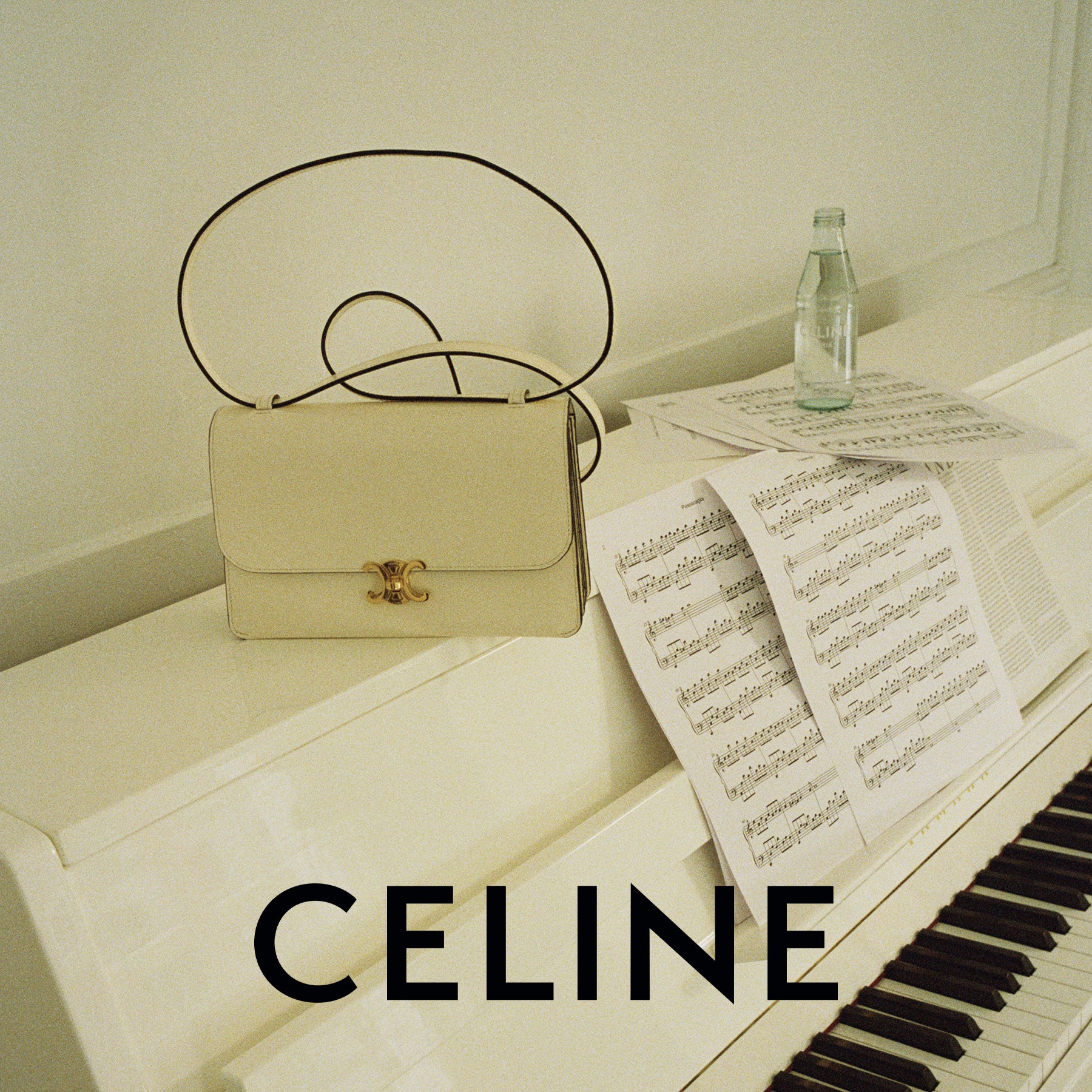 A Celine shoulder bag in light beige with a gold interlocking CC clasp and a thin black strap, styled on a white piano next to sheet music and a glass bottle, with the word CELINE in bold text at the bottom.