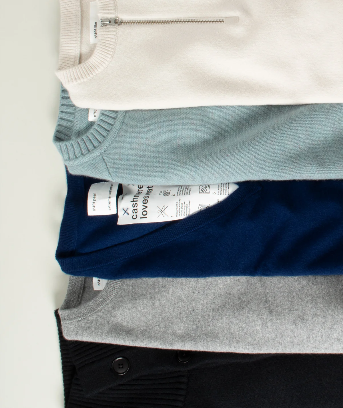 Four folded knit crewneck sweaters stacked vertically, showing different colors: off-white, light blue, navy blue, and heather gray, with a black sweater partially visible at the bottom all in Extreme Cashmere brand. 