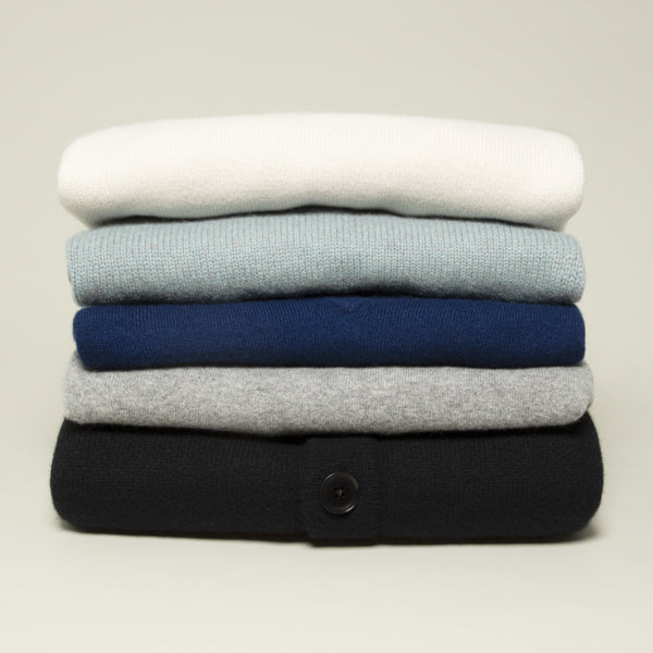 A stack of five folded knit sweaters in white, light blue, navy, heather gray, and black, arranged vertically on a light beige background all in Extreme Cashmere.