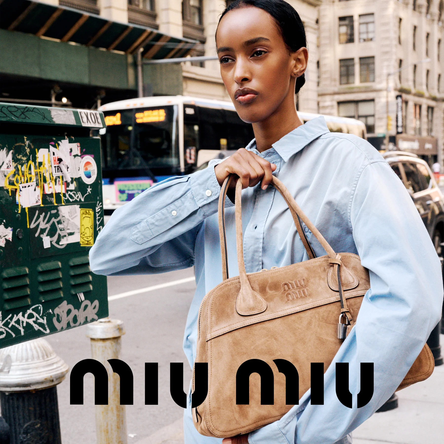 A model in a light blue denim shirt holds a tan Miu Miu suede briefcase bag on a city street. The Miu Miu brand name is prominently displayed in black text at the bottom of the image.