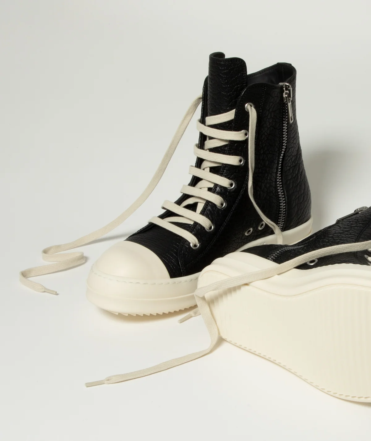 A pair of Rick Owens black high-top sneakers with textured leather uppers, cream laces, and thick cream platform soles, shown on a light gray studio background. 