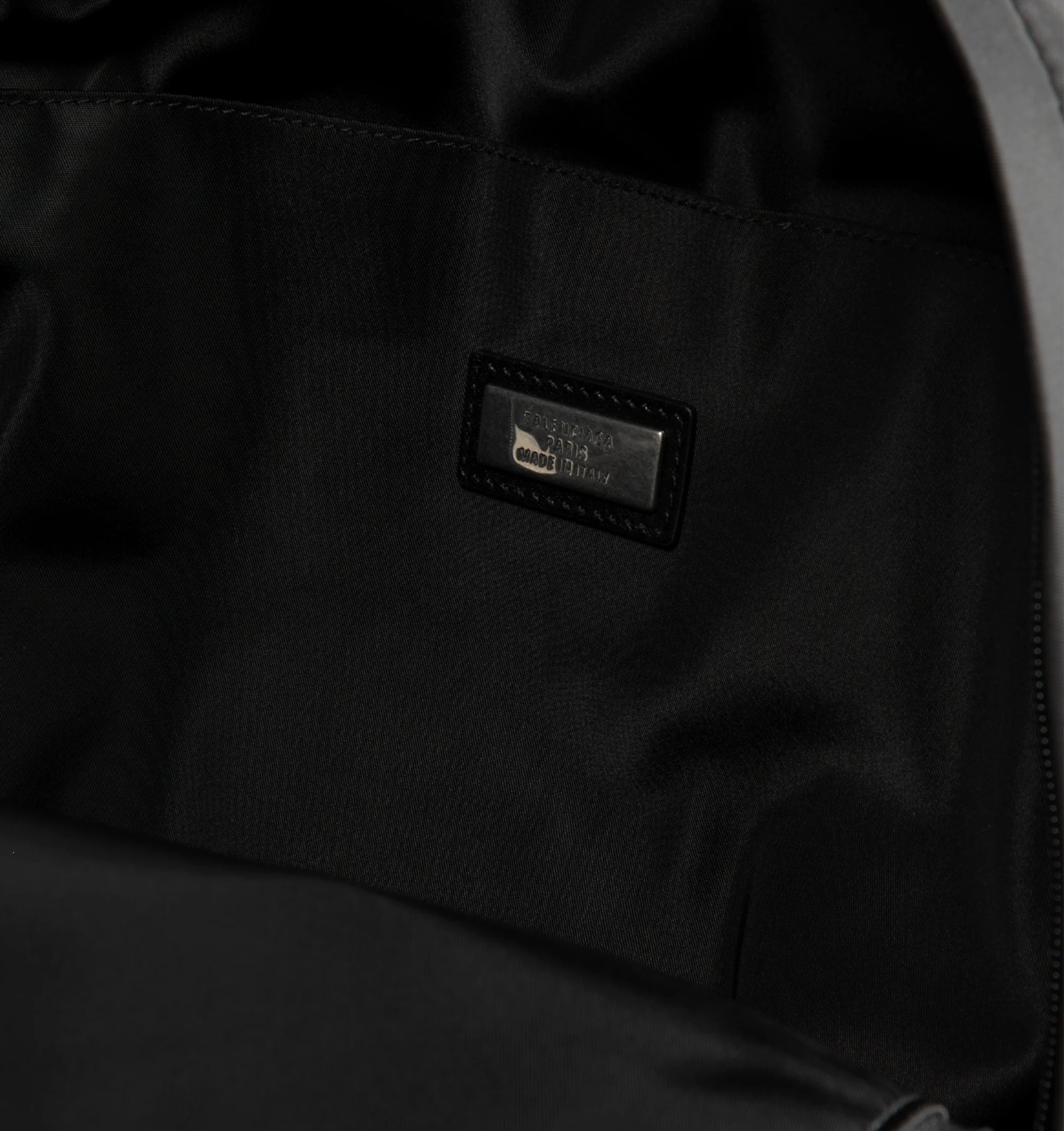 Image 3 of 3 - BLACK - Balenciaga Sleek Mens Nylon Backpack is crafted from black recycled nylon with smooth leather trim and features a zip-around closure and adjustable padded shoulder straps with metal hardware. A circular top handle, front logo print, metal logo plaque, interior pocket, and front compartment with two zippers complete the design. Fits a tablet, notebook, wallet, phone, keys, and small water bottle. Main Material: 100% Recycled Nylon. Leather Detail: Smooth Leather. Dimensions: 30 x 14 x