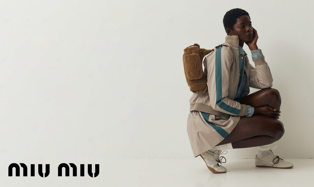 Person wearing Miu Miu beige and blue jacket with a brown bag, sitting on the floor against a white background. 