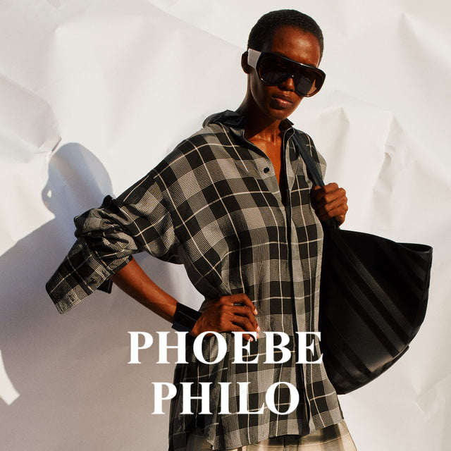 Person wearing a plaid shirt and sunglasses, holding a black bag with 'Phoebe Philo' branding.